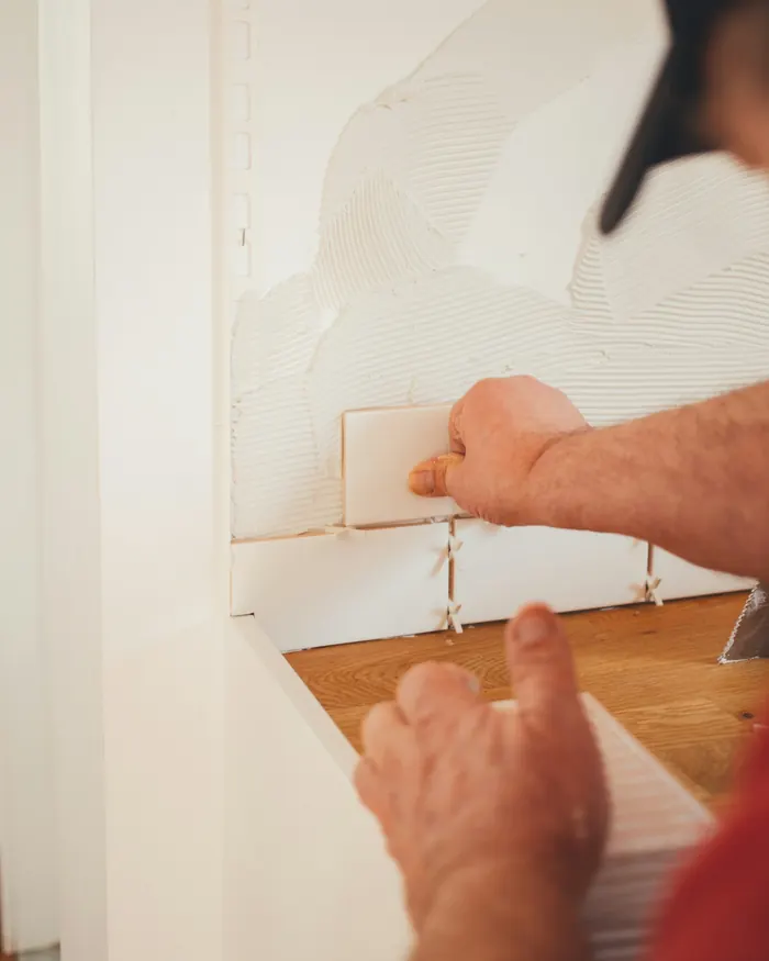 Tiler pressing a white wall tile into fresh adhesive, with spacers in the joints below